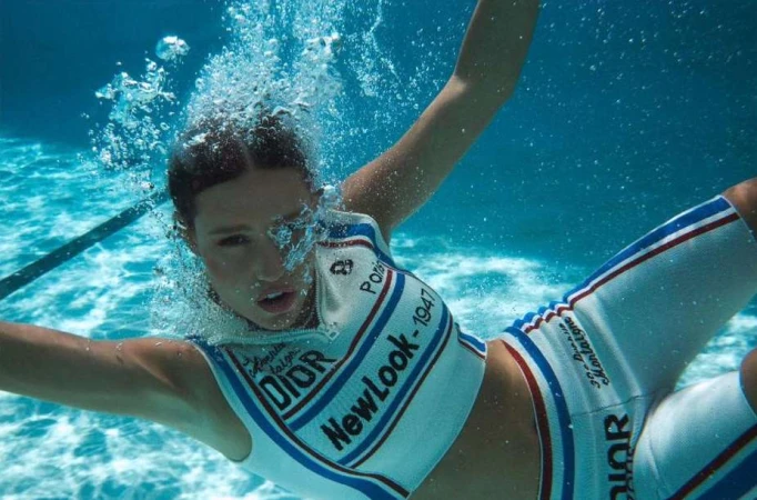 Underwater shot of a woman wearing a white and blue Dior-branded outfit, surrounded by bubbles in a bright, clear blue pool.