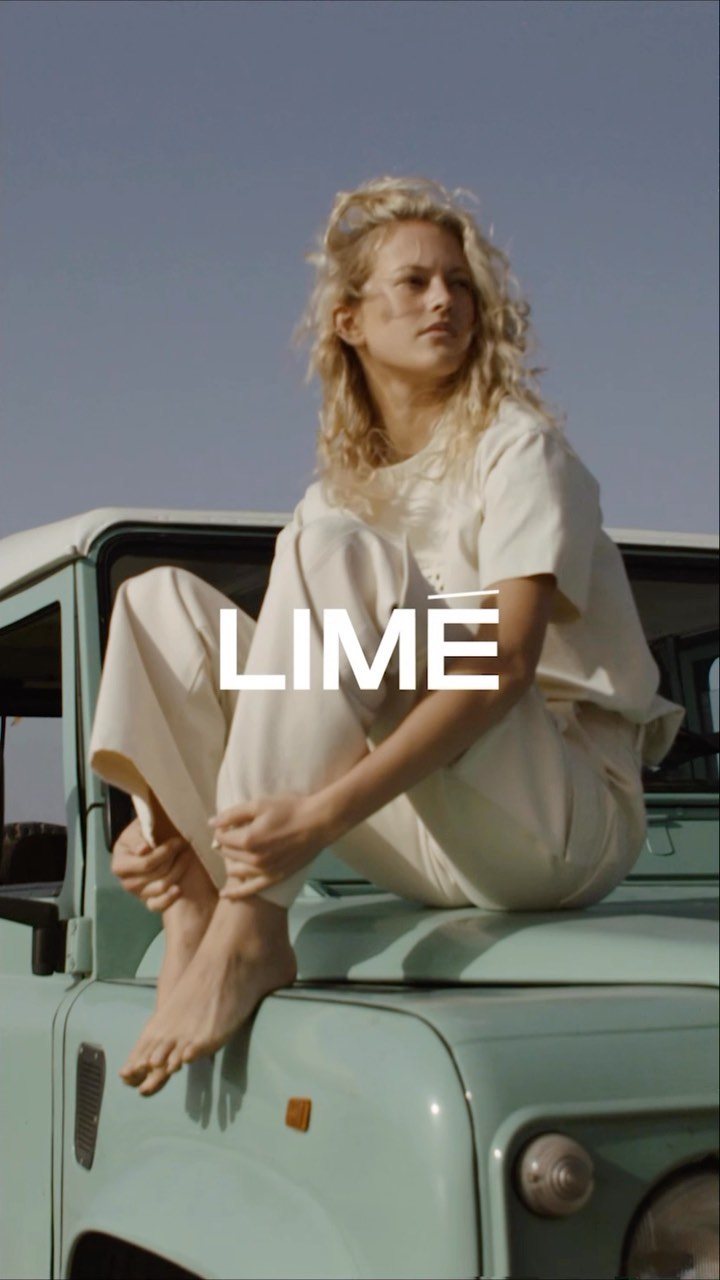A woman with curly blonde hair in cream-colored casual clothing sits on the hood of a light green SUV in an outdoor, desert-like setting.