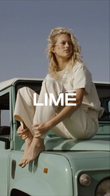 A woman with curly blonde hair in cream-colored casual clothing sits on the hood of a light green SUV in an outdoor, desert-like setting.