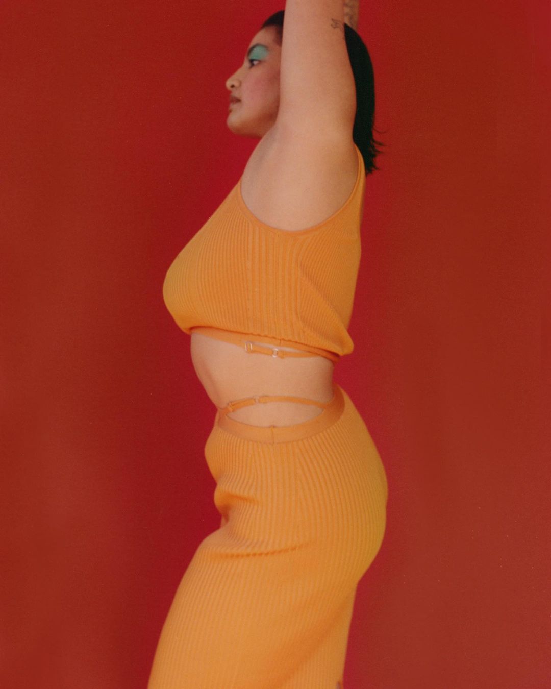 A woman in a profile shot is wearing a matching knit top and skirt, posing against a vibrant red backdrop with striking makeup.