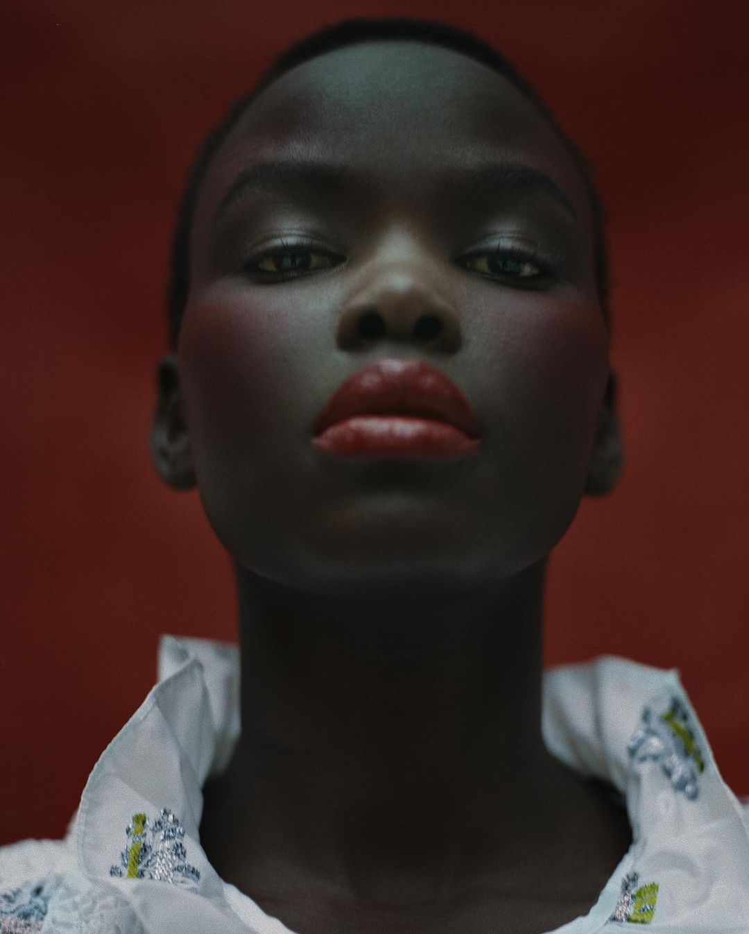 Close-up portrait of a Black woman with short hair, bold red lipstick, and a white embroidered high-collar top against a deep red background.