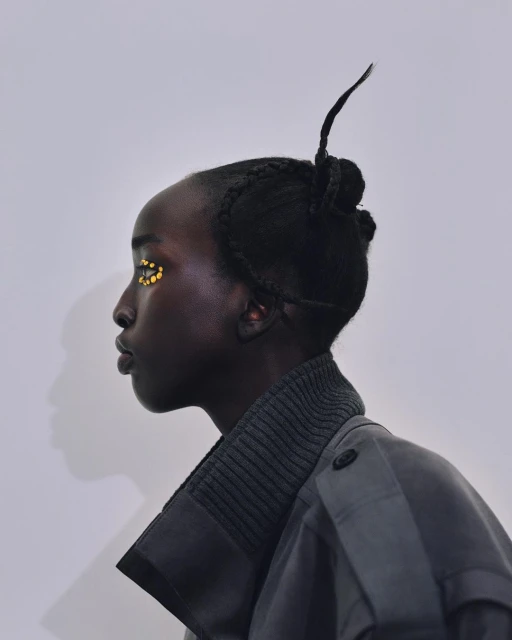 Profile portrait of a Black woman with a unique braided hairstyle, wearing a gray coat with golden accents near her eye against a gray background.
