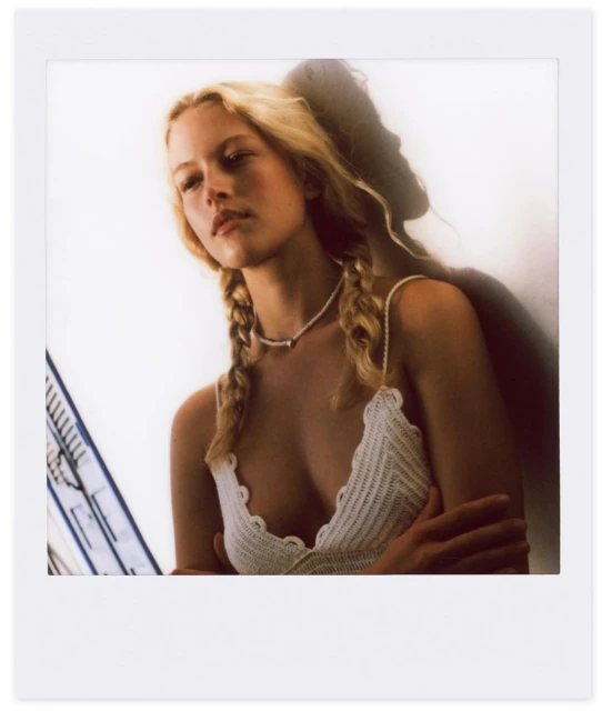 A woman with blonde hair in pigtails poses with her arm around another person in a soft-lit, intimate portrait on white background.
