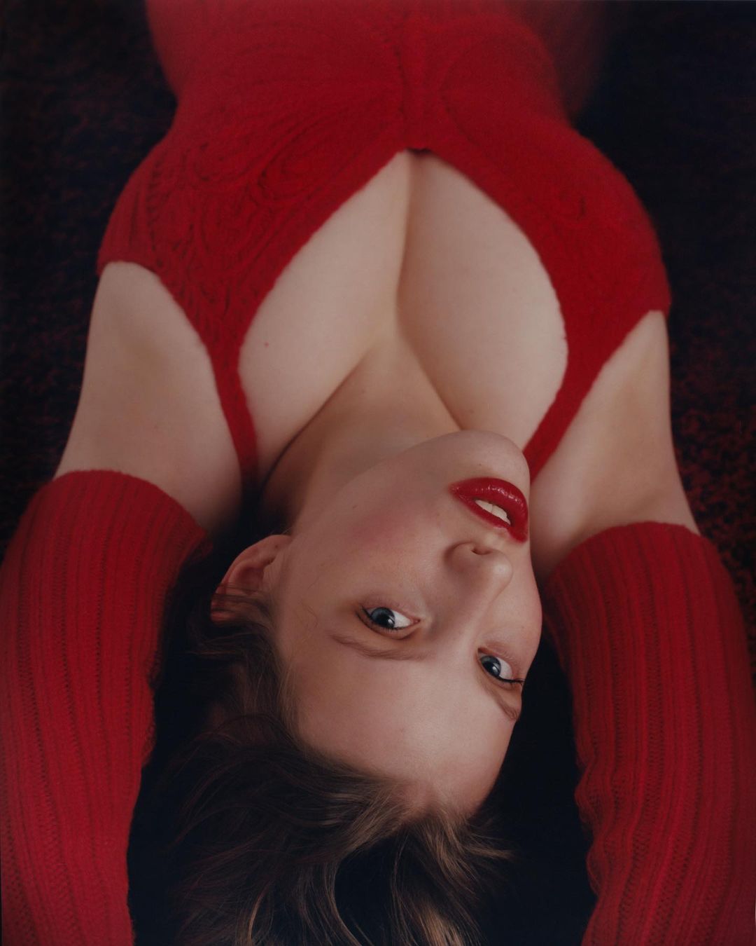 Portrait of a blonde woman in a red knit dress lying on her back looking at the camera, styled with red lipstick. 