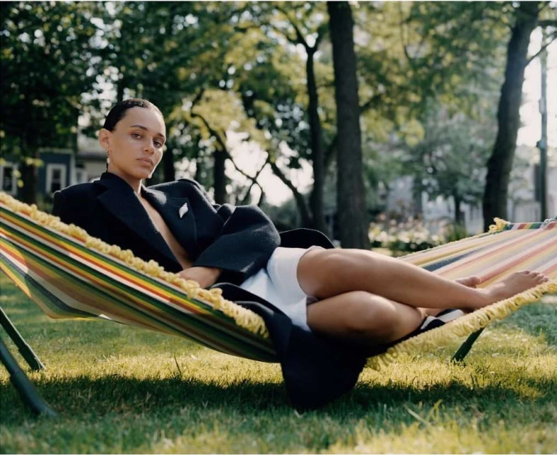 Adult woman relaxes in a colorful hammock on a lawn surrounded by green trees in a residential area. She wears a black blazer and white shorts.