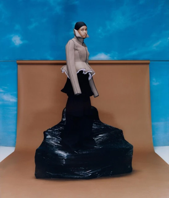Fashionable woman in beige peplum jacket posing in studio against a blue cloudy backdrop, with black plastic wrapped around her lower body.