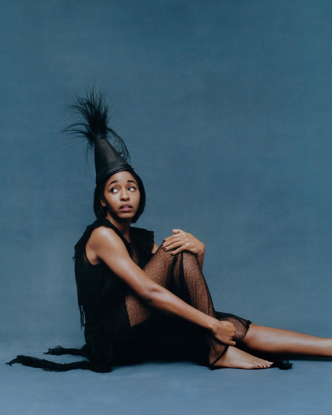 Seated black woman wearing a dark sheer dress and feathered conical hat against a textured blue backdrop
