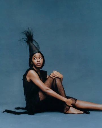 Seated black woman wearing a dark sheer dress and feathered conical hat against a textured blue backdrop