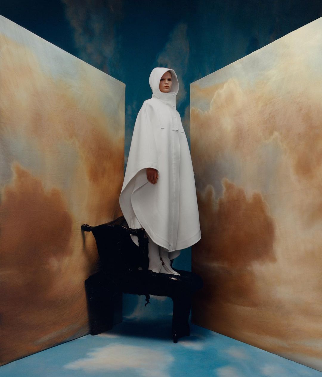 Fashion photography of a person wearing a hooded poncho, posing atop a black sculptural chair inside a studio with a painted sky backdrop.