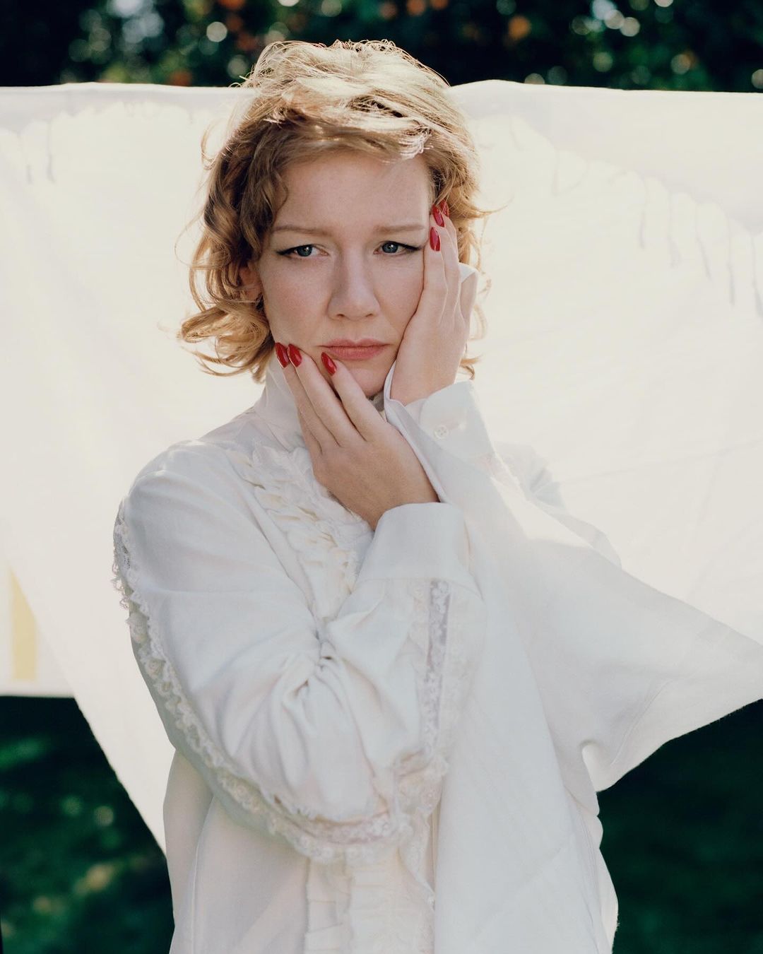 Adult female model with red nails poses in front of a white sheet outdoors, wearing a white blouse with lace details, captured in natural lighting.