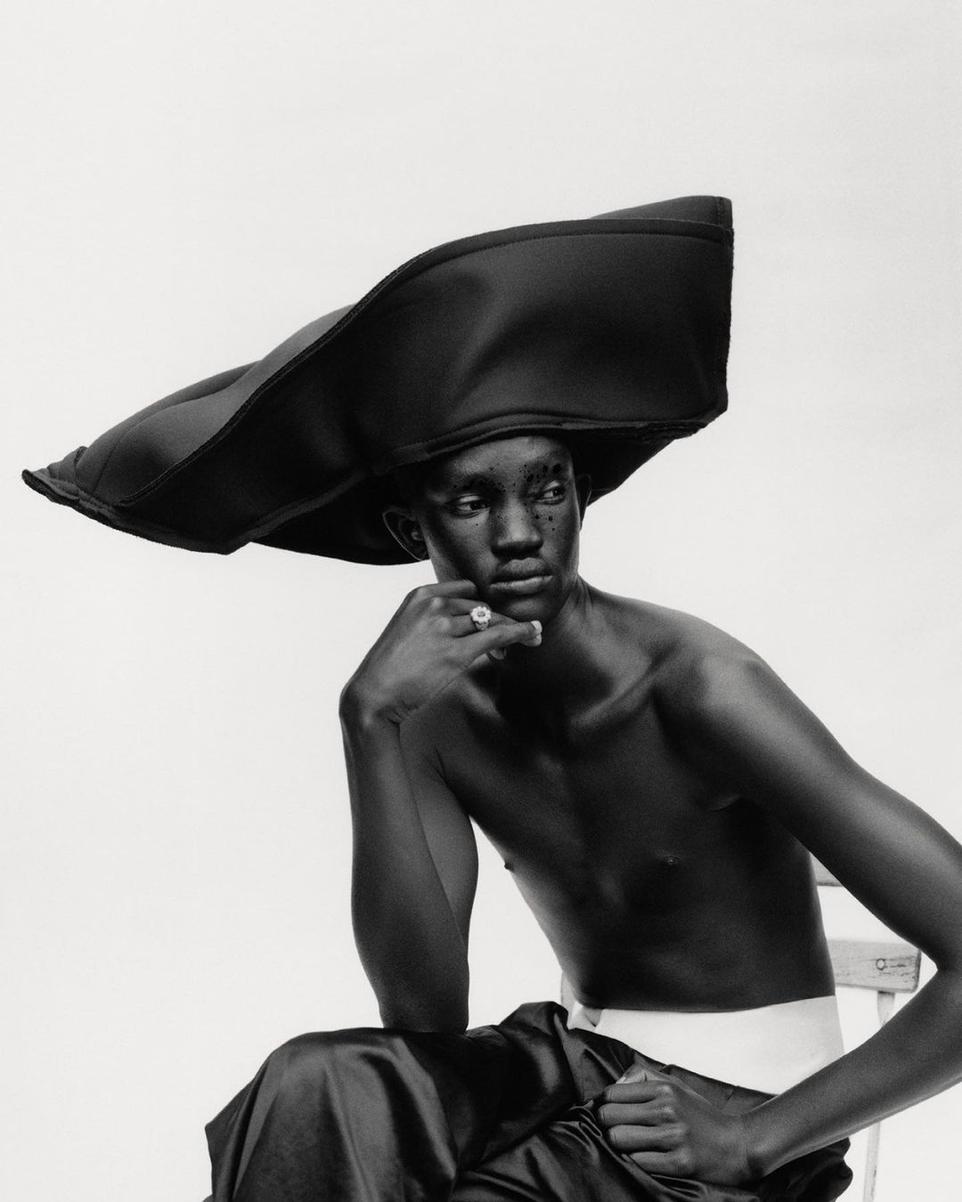 Black and white studio portrait of an African male fashion model wearing a large brimmed hat while seated on a chair.
