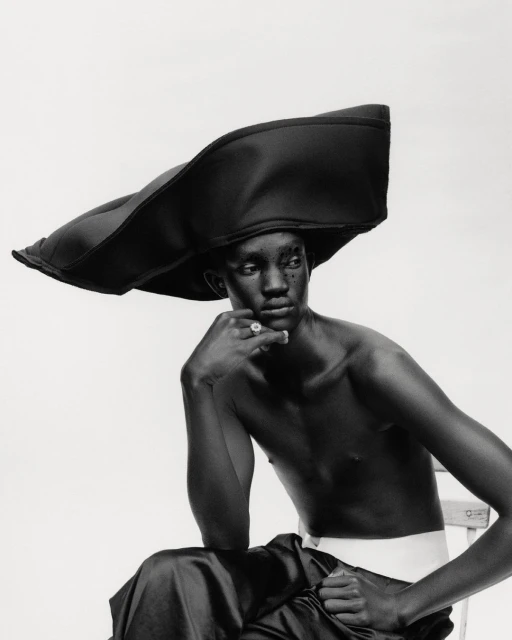 Black and white studio portrait of an African male fashion model wearing a large brimmed hat while seated on a chair.