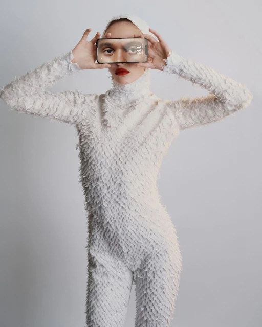A woman in a textured white outfit holds a smartphone up to her face, obscuring her eyes. She has red lipstick and a neutral expression.