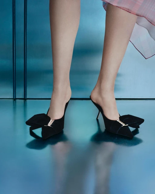 Close-up of woman's legs and feet in black high-heeled mule shoes with embellished bow detail against blue reflective backdrop.