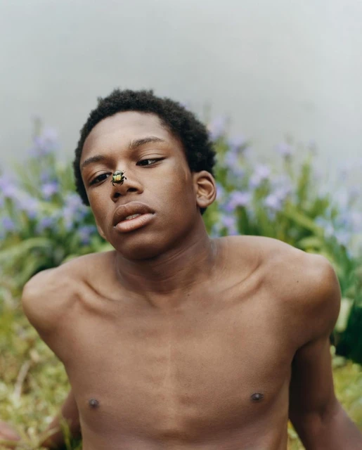 Portrait of a young Black man with a beetle on his nose, captured outdoors with soft, natural light and a background of purple flowers.