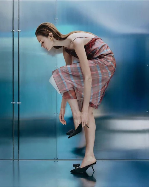 A slender woman bends over, putting on a black mule in a fashion shot. She wears a plaid dress, indoors with blue background.