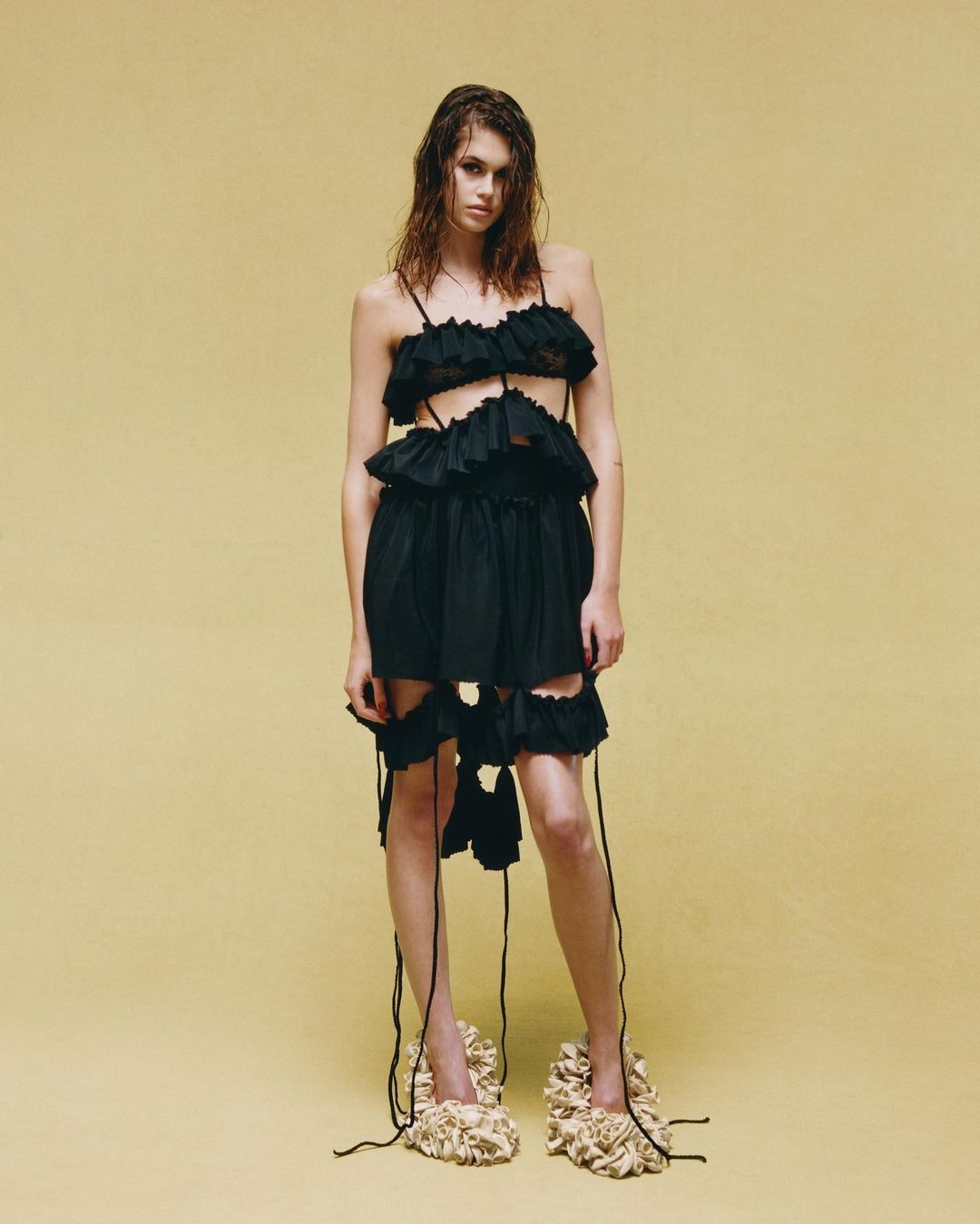 Full-length shot of a woman in a black, tattered dress with wet hair, standing in front of a yellow seamless backdrop in a studio.
