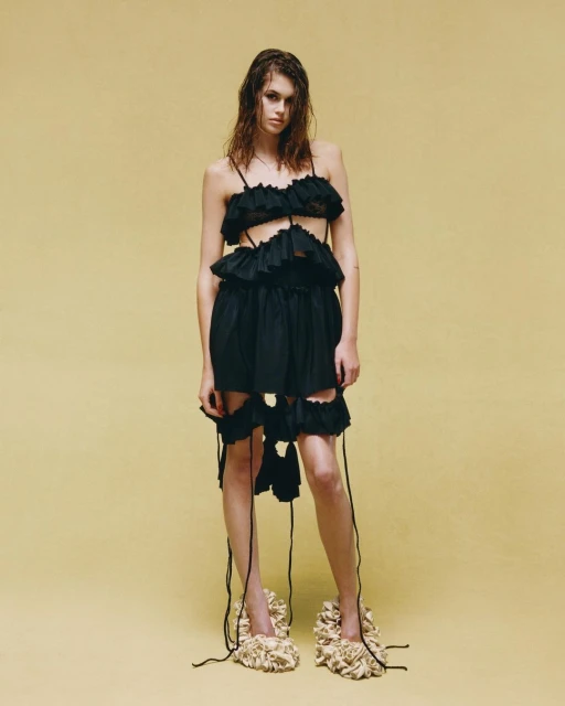 Full-length shot of a woman in a black, tattered dress with wet hair, standing in front of a yellow seamless backdrop in a studio.