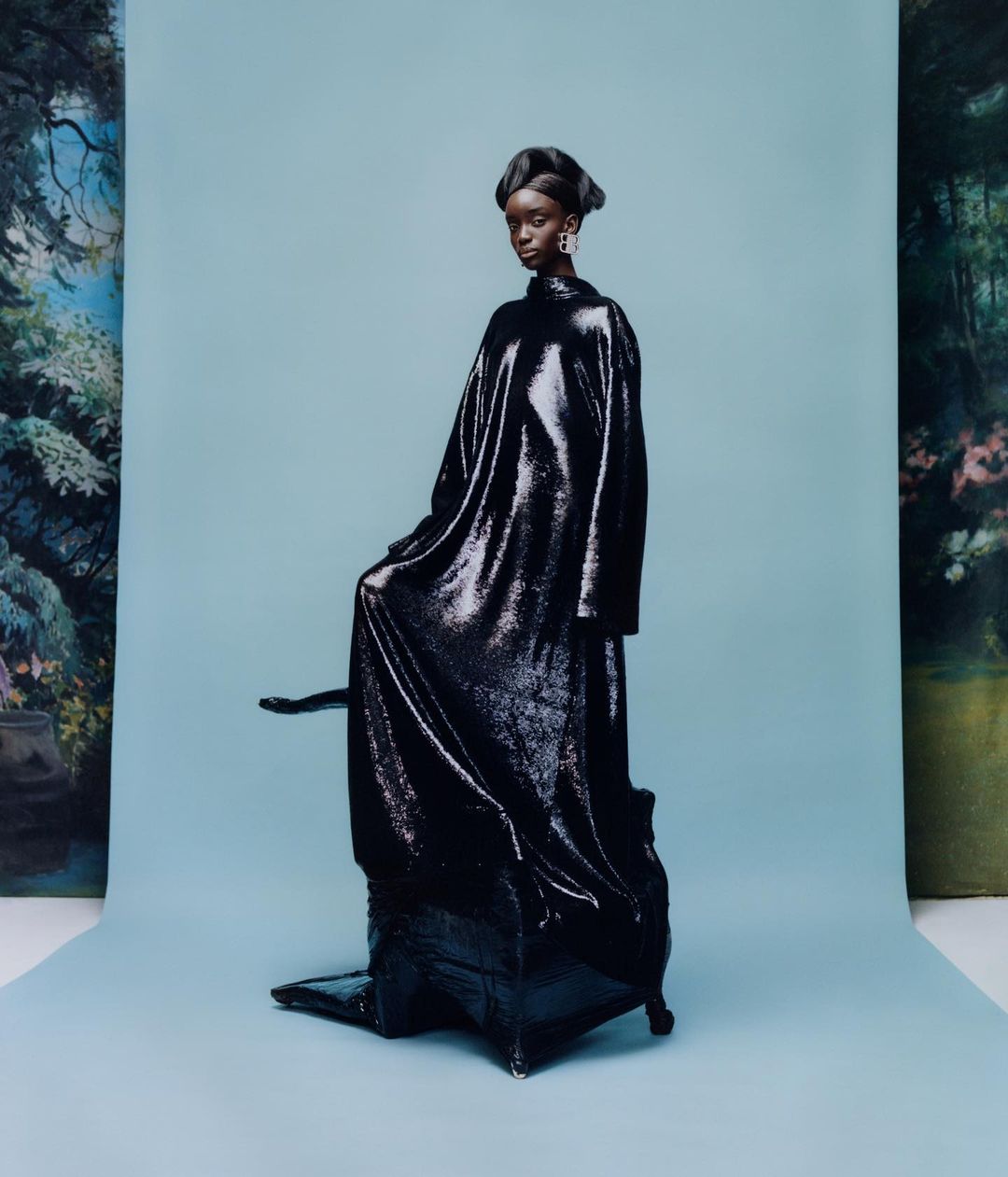 A black female fashion model poses in an oversized, long-sleeved, sequined dress against a pastel blue backdrop with a painted forest scene.