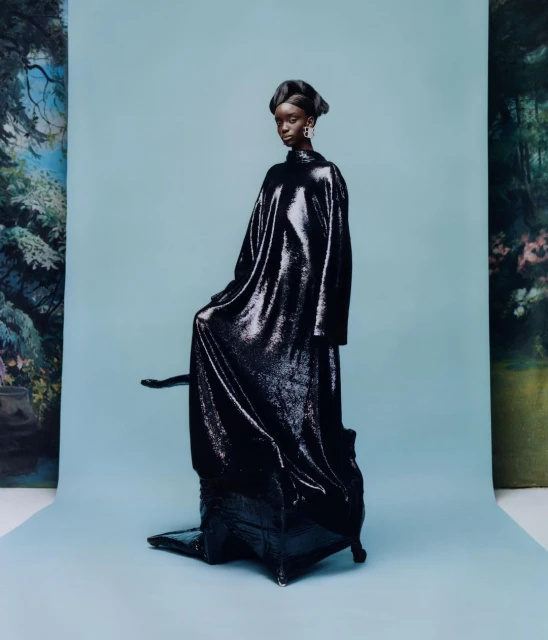 A black female fashion model poses in an oversized, long-sleeved, sequined dress against a pastel blue backdrop with a painted forest scene.