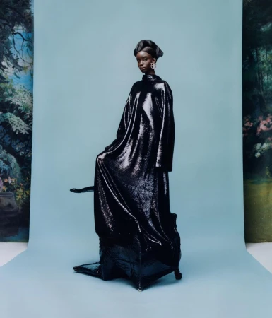 A black female fashion model poses in an oversized, long-sleeved, sequined dress against a pastel blue backdrop with a painted forest scene.