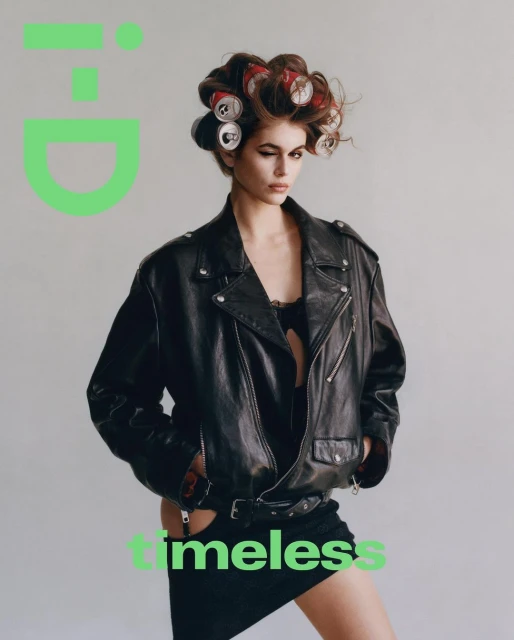 Fashion editorial shot of a model with curlers, wearing a leather jacket in front of a solid gray backdrop, posing for a magazine cover.