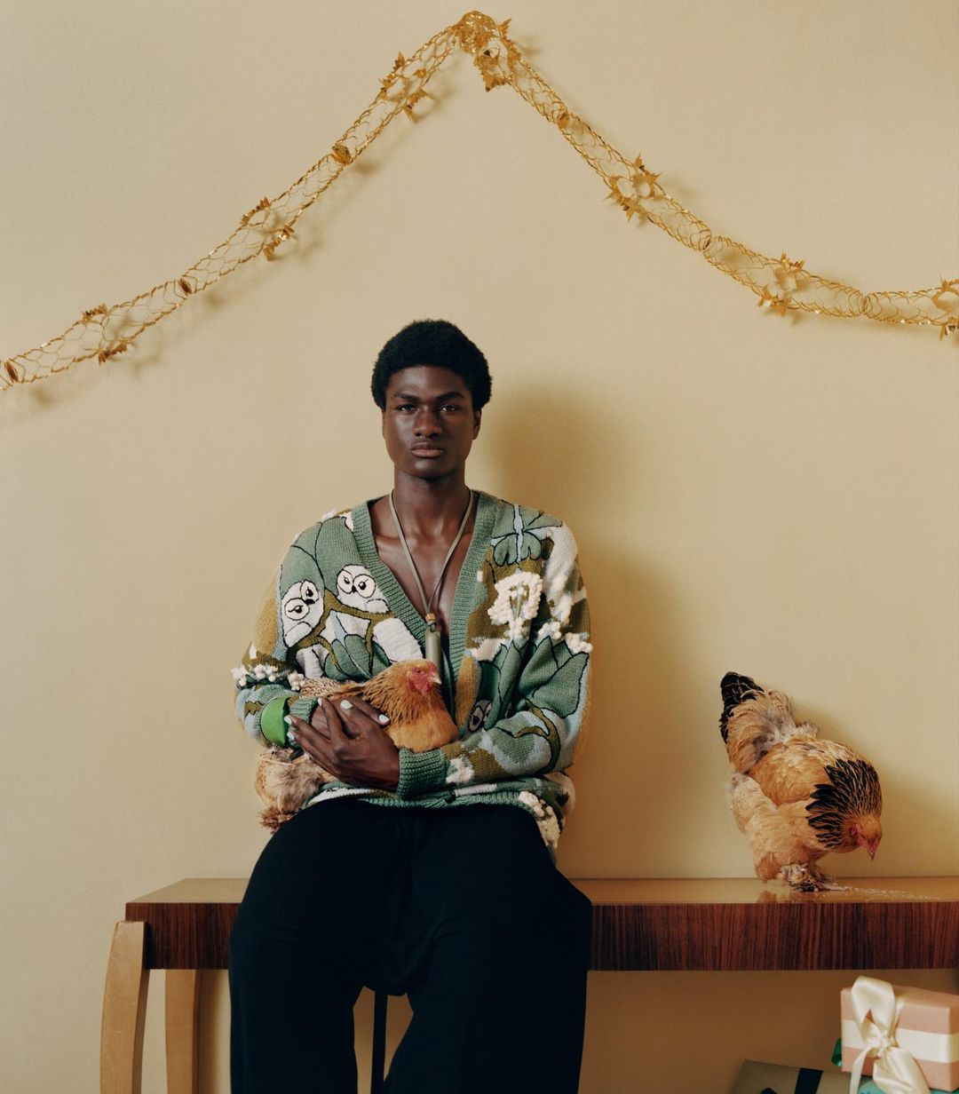 Portrait of a Black male model in patterned cardigan, holding a chicken, with another chicken beside him on a wooden table, gifts below.