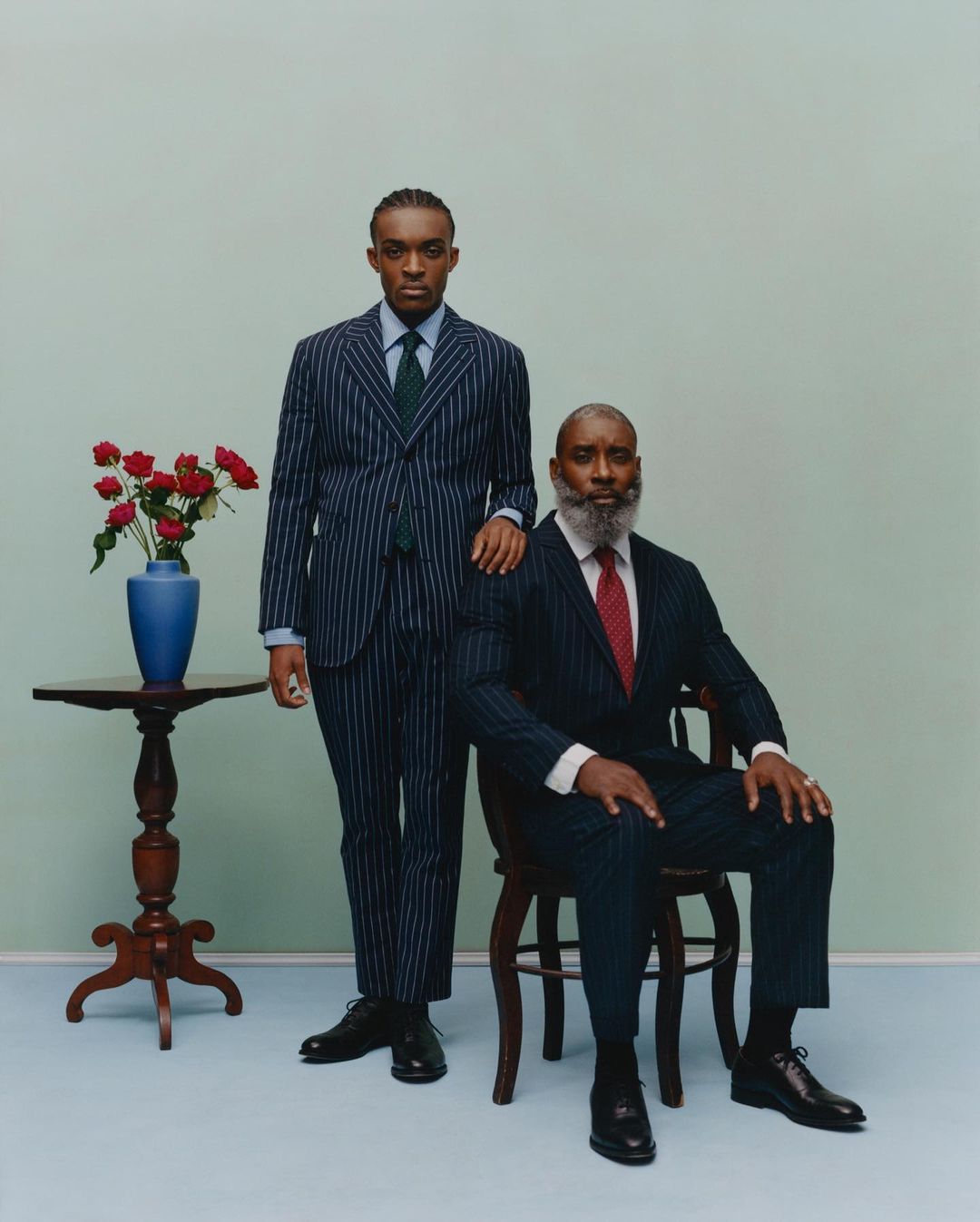 Two formally dressed men in suits pose indoors next to a table with red roses in a blue vase. One man sits on a wooden chair.