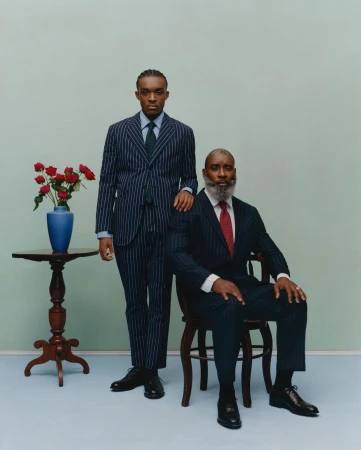 Two formally dressed men in suits pose indoors next to a table with red roses in a blue vase. One man sits on a wooden chair.