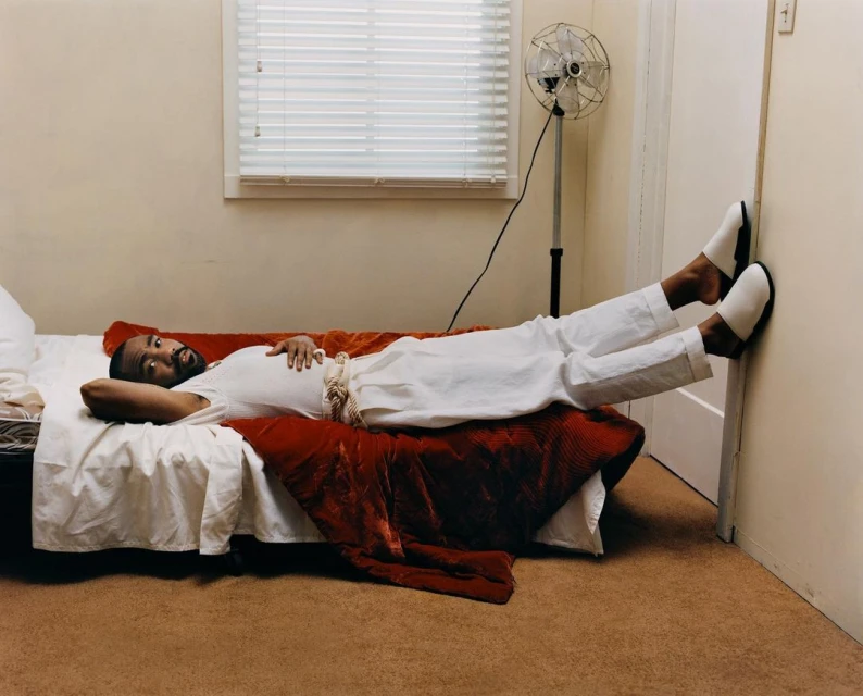 An adult man lies on a bed with his legs propped against a wall, wearing white pants and slippers in a brightly lit room.