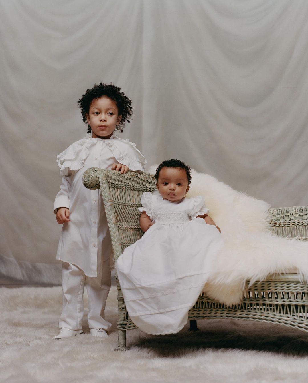 Two young children, one standing and one seated on a wicker chaise lounge covered in a white fur throw, pose in a neutral indoor setting.