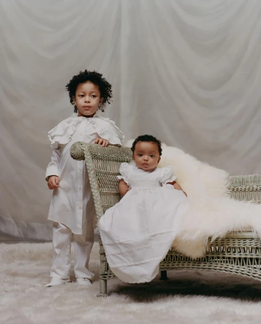 Two young children, one standing and one seated on a wicker chaise lounge covered in a white fur throw, pose in a neutral indoor setting.