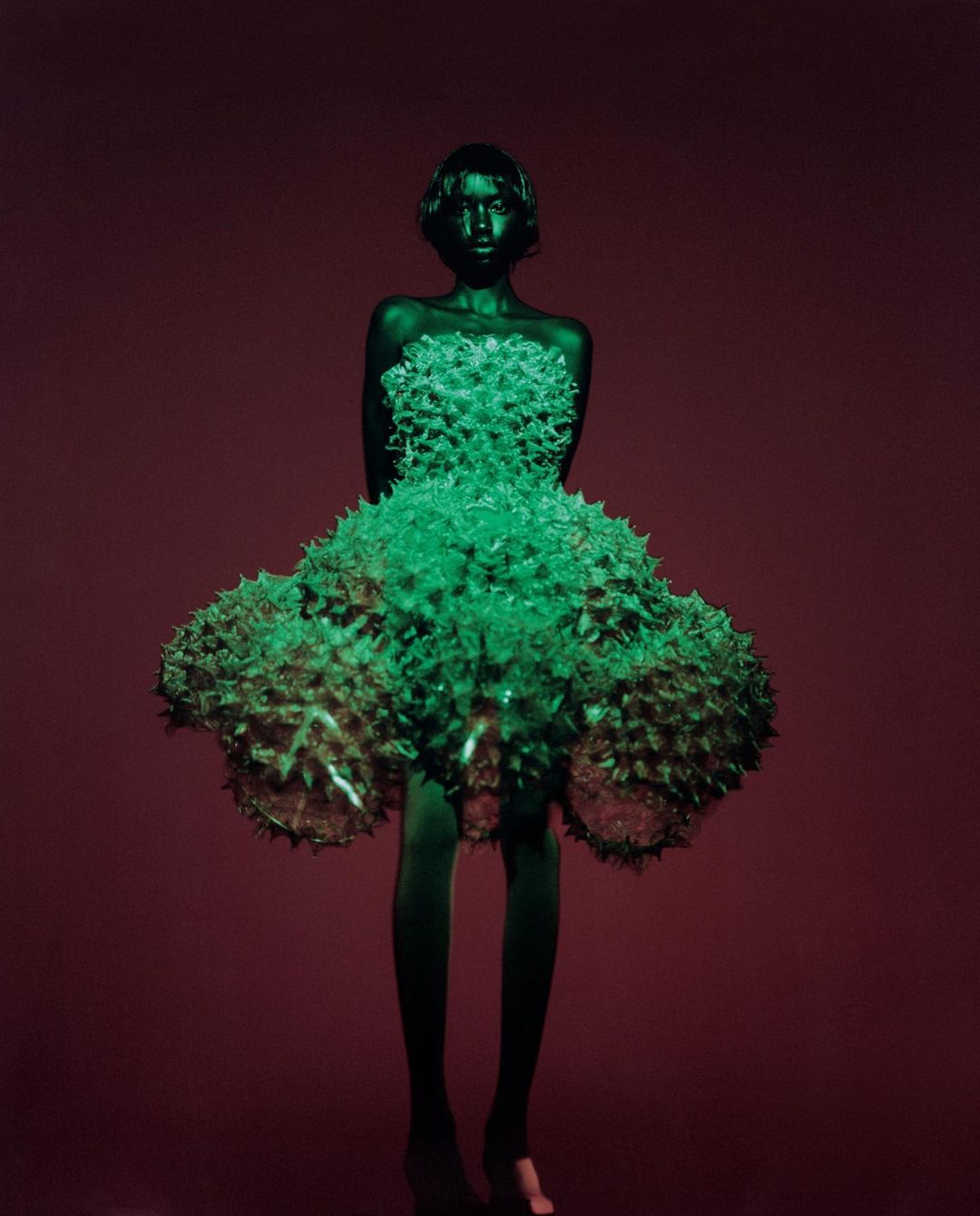 Full shot of a model wearing a green spiky gown against a deep red backdrop, in a studio setting with bright, contrasting lighting.
