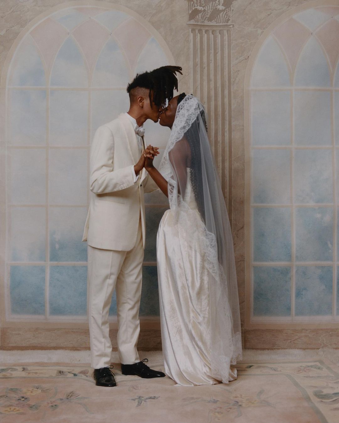 A bride and groom in wedding attire share a kiss in front of a painted backdrop featuring arched windows and classical architectural elements.