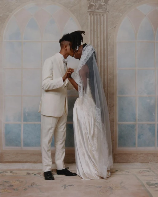 A bride and groom in wedding attire share a kiss in front of a painted backdrop featuring arched windows and classical architectural elements.