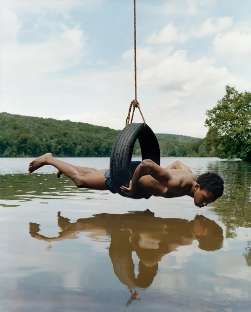 Young man hangs from a tire swing over a lake, reflected in the water, surrounded by lush greenery and a cloudy sky in daylight.
