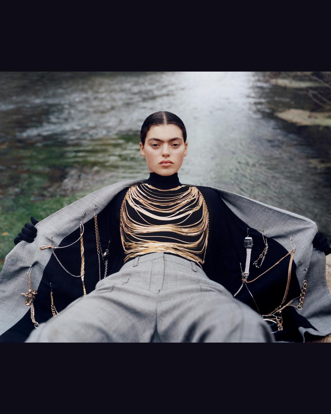 Woman in a black turtleneck and grey trousers poses with a grey blazer in an outdoor river setting, showcasing gold jewelry.
