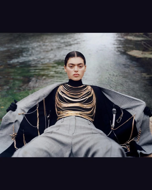 Woman in a black turtleneck and grey trousers poses with a grey blazer in an outdoor river setting, showcasing gold jewelry.