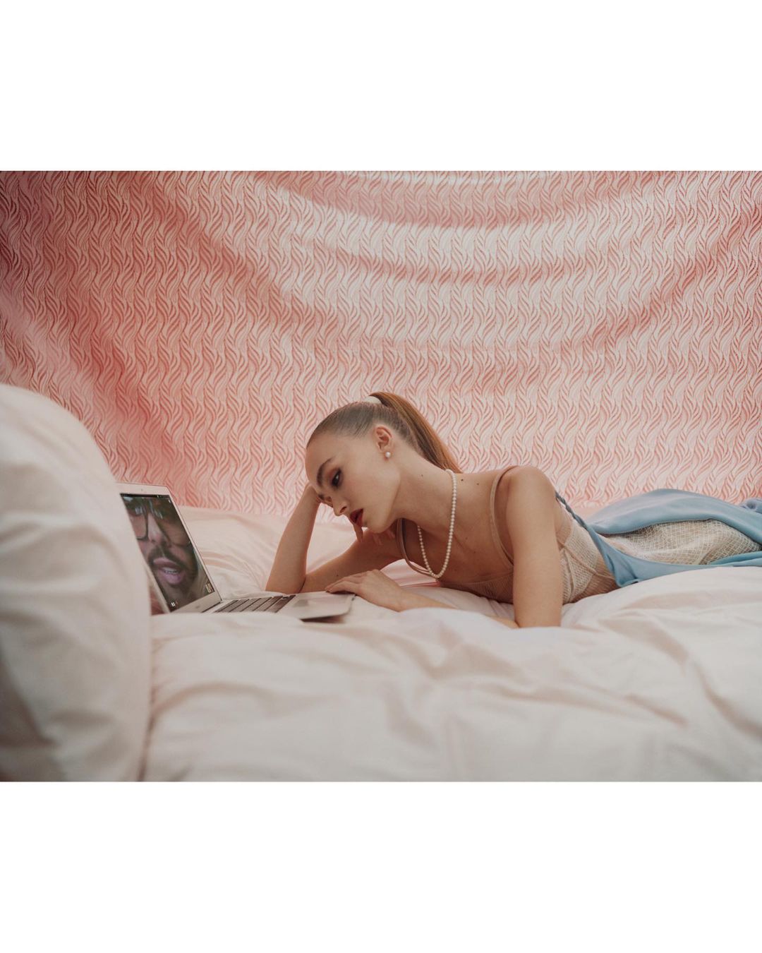 A young fair-skinned woman with a ponytail and pearl necklace lies on a bed, looking at a laptop in a brightly-lit indoor bedroom setting.