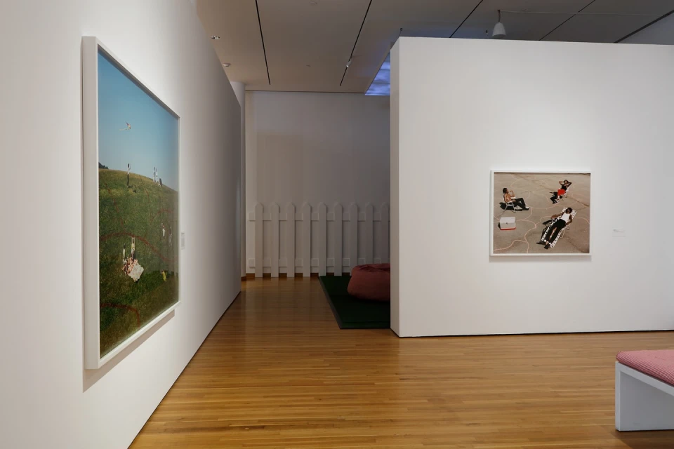 Interior of an art gallery featuring framed photographs on white walls and a wooden floor, with a white picket fence and beanbag in the background.