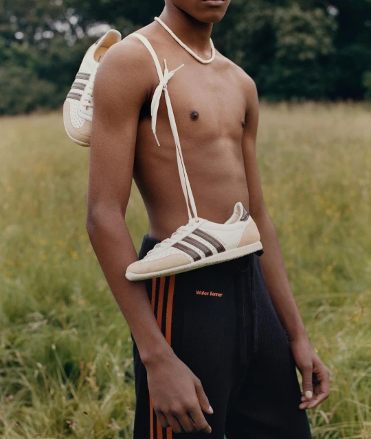 Athletic man wears Adidas Samba sneakers like a bag, outdoors on a green field, with a black track suit and a white necklace.