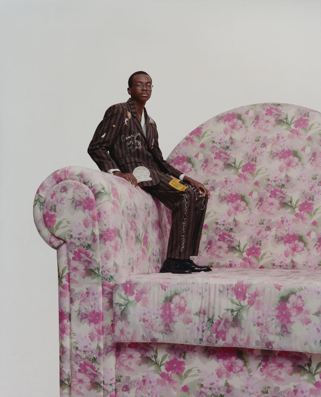 Young man in a striped suit with floral sofa. He is seated, looking at the camera against a plain background. The sofa has a pink floral pattern.