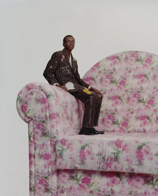 Young man in a striped suit with floral sofa. He is seated, looking at the camera against a plain background. The sofa has a pink floral pattern.