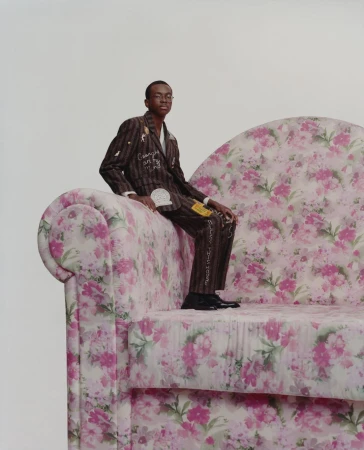 Young man in a striped suit with floral sofa. He is seated, looking at the camera against a plain background. The sofa has a pink floral pattern.