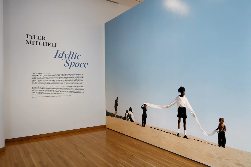 Tyler Mitchell's artwork 'Idyllic Space' featuring people on a sandy surface with a light blue sky, exhibited in an art gallery.