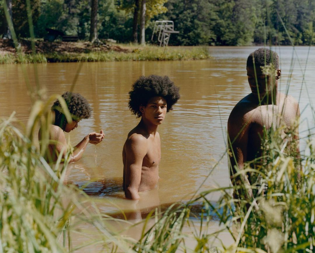 Three topless young adult men stand waist-deep in a brown lake, framed by tall green grasses and trees with a blue diving platform.