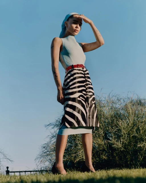 Elegant adult woman in zebra-print skirt, headscarf, and heels standing outside under the sunlight against a clear blue sky.