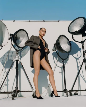 A woman in a black bodysuit and gray blazer poses among studio lights against a white background, capturing a modern and chic fashion aesthetic.