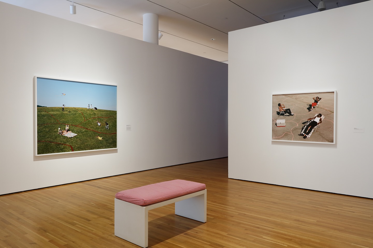Interior view of an art gallery with a wooden bench, framed photographs of people outdoors, and white walls on a hardwood floor.