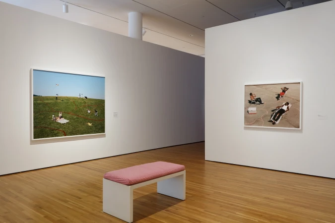 Interior view of an art gallery with a wooden bench, framed photographs of people outdoors, and white walls on a hardwood floor.